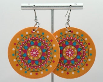 May include: Two circular earrings with an orange base and a colourful mandala pattern. The mandala design includes pink, teal, and yellow details. The earrings are presented on a silver stand against a white backdrop.