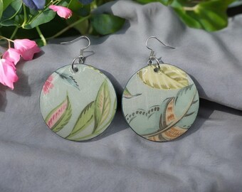 May include: A pair of circular earrings with a floral pattern. The earrings display a pale blue backdrop adorned with green leaves, pink flowers, and other botanical motifs. They are fitted with silver-coloured hooks.