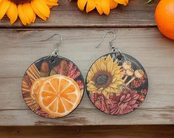 May include: Circular earrings with autumnal designs. One earring displays a sunflower and flowers, the other features orange slices, cinnamon sticks, and acorns. Both have silver hooks and a dark background.