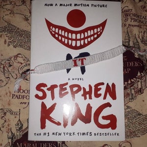 May include: A copy of Stephen King's novel "IT" with a white and red bracelet laid across the cover. The book cover features a clown face and the title in large red letters. The background is a patterned fabric.