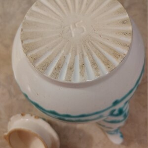 Pu&ograve; includere: Teiera in ceramica bianca con un designo verde e bianco. La teiera ha un coperchio con un designo circolare e il numero "45" stampato sul fondo. La teiera ha tre gambe e un piccolo coperchio.