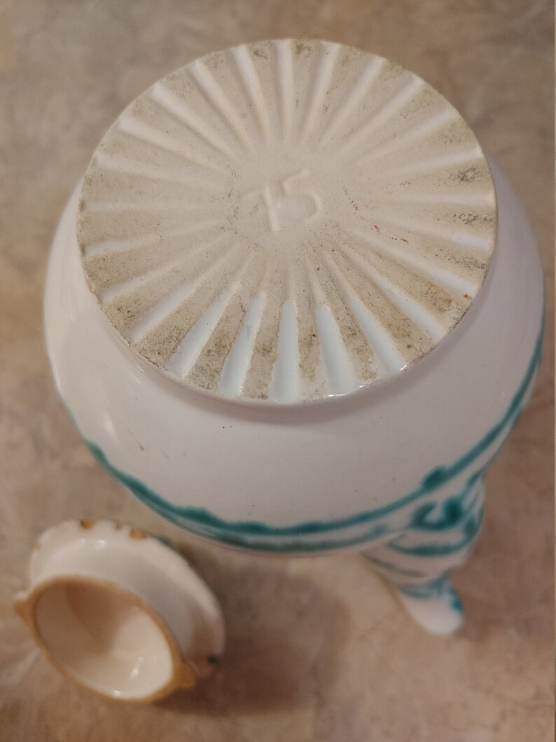 Pu&ograve; includere: Teiera in ceramica bianca con un designo verde e bianco. La teiera ha un coperchio con un designo circolare e il numero "45" stampato sul fondo. La teiera ha tre gambe e un piccolo coperchio.