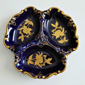 May include: A three-sectioned, navy blue ceramic serving dish with gold floral designs and trim. The dish has a scalloped edge and is likely used for serving appetizers or small portions of food. The gold accents provide an elegant contrast to the dark blue.