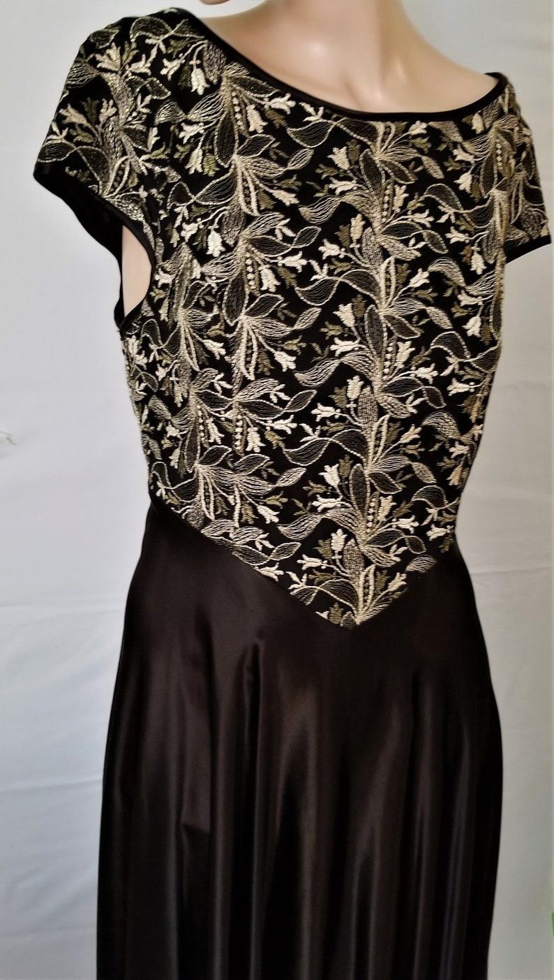 以下が含まれることがあります： ゴールドとグリーンの糸で花柄の刺繍が施された、黒いサテンのドレス。ドレスはスクープネックと短い袖です。