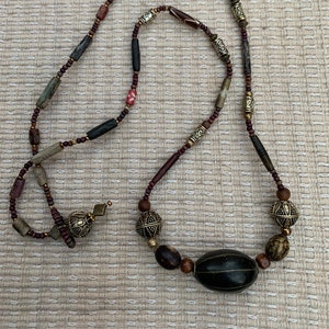 May include: A long necklace made of various beads, including wood, metal, and stone. The necklace features a large, dark brown, oval-shaped bead in the center.