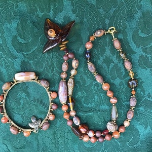 May include: A set of two necklaces made with brown, orange, and gold beads. The necklace on the left is a bracelet with a gold wire wrapped around the beads. The necklace on the right is longer and has a gold clasp. Both necklaces feature a variety of natural stone beads.