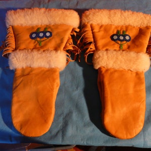 May include: A pair of orange leather mittens with white fur trim and beaded floral designs. The cuffs have fringe and are lined with white fur. These mittens are designed for warmth and protection from the cold.