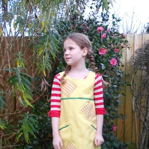 May include: A young girl wearing a yellow dress with red and white stripes and a green sash. The dress has a diamond pattern and pockets. She is standing in front of a green bush.