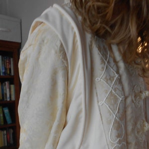 Puede incluir: Una prenda blanca de manga larga con un ribete de encaje decorativo y una tela texturizada. La prenda tiene un cuello alto y un corpiño ajustado.