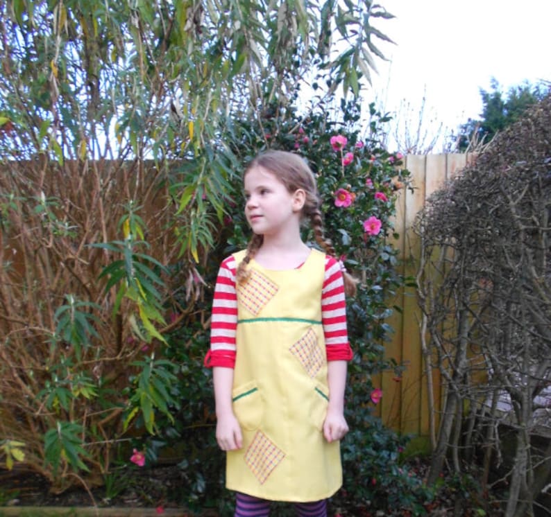May include: A young girl wearing a yellow dress with red and white stripes and a green sash. The dress has a diamond pattern and pockets. She is standing in front of a green bush.