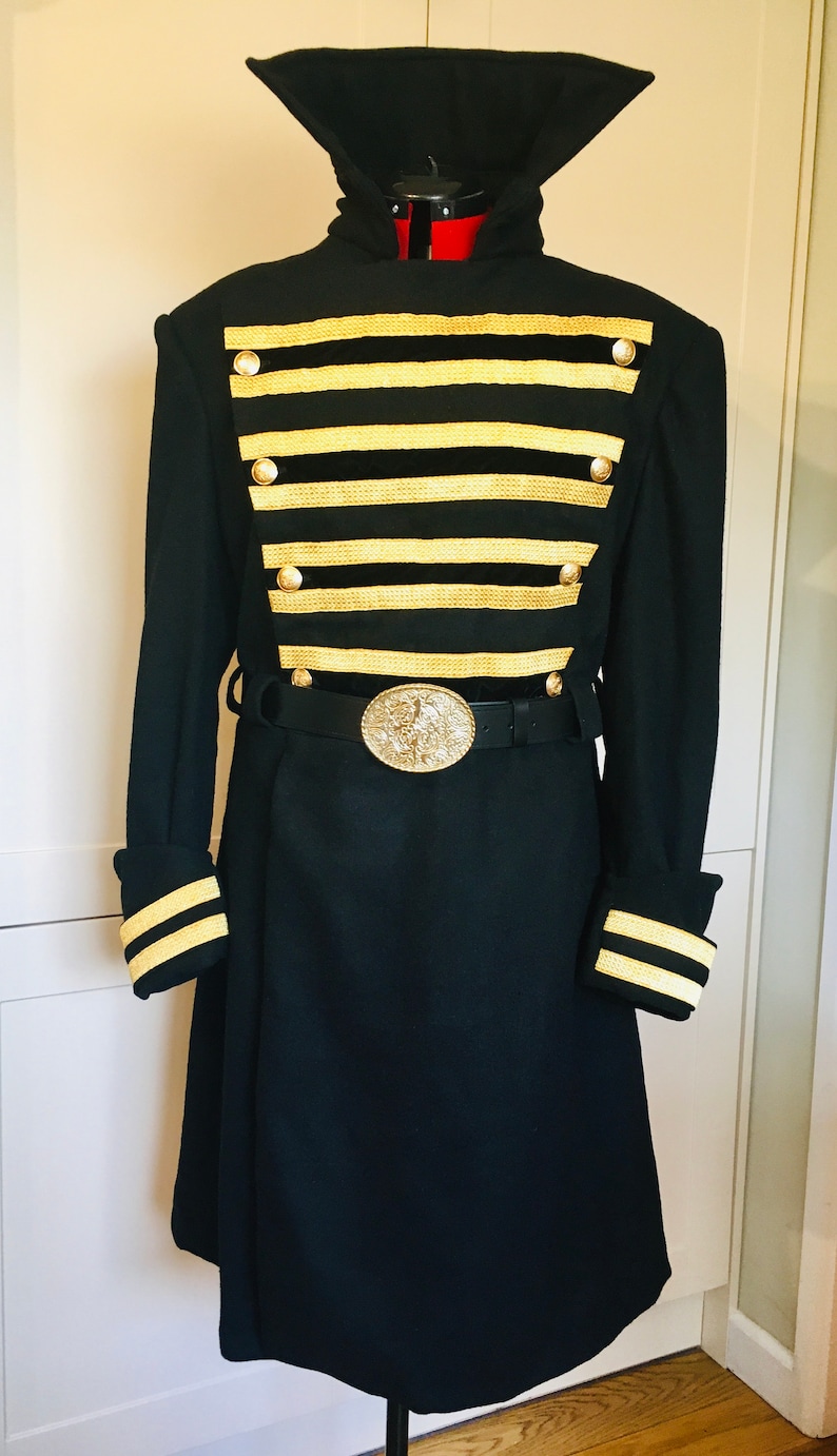 May include: A black long coat with a high collar and gold trim. The coat has a belt with a large, ornate buckle. The coat is on a mannequin.