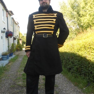 May include: A man wearing a black frock coat with gold and black striped trim. The coat has a belt and is worn over a white shirt. The man is standing on a gravel path.