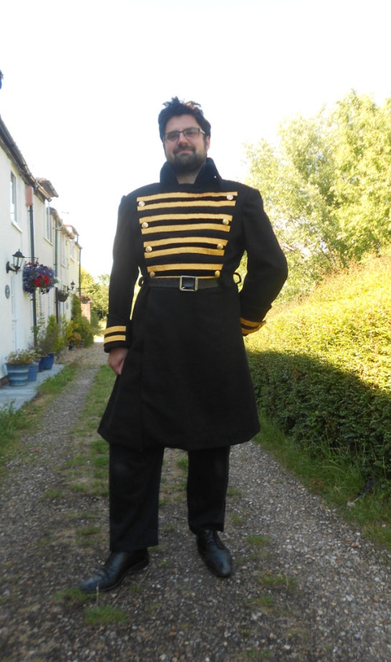May include: A man wearing a black frock coat with gold and black striped trim. The coat has a belt and is worn over a white shirt. The man is standing on a gravel path.