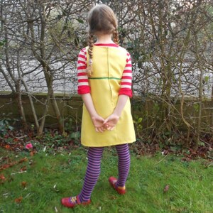 May include: A young person wearing a yellow dress with red and white stripes on the sleeves, purple and white striped tights, and red shoes. The dress has a green belt and a zipper down the back.