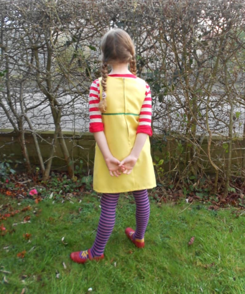 May include: A young person wearing a yellow dress with red and white stripes on the sleeves, purple and white striped tights, and red shoes. The dress has a green belt and a zipper down the back.