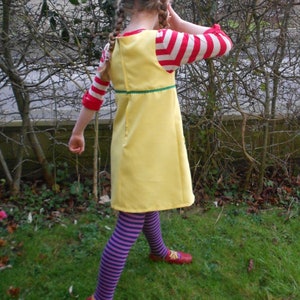 May include: A young girl wearing a yellow dress with a green trim, red and white striped sleeves, purple and white striped tights, and red shoes. She is standing in a grassy area with trees in the background.