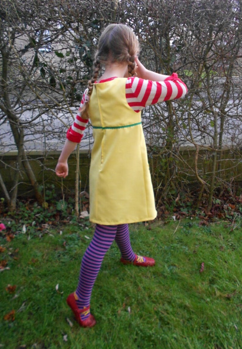 May include: A young girl wearing a yellow dress with a green trim, red and white striped sleeves, purple and white striped tights, and red shoes. She is standing in a grassy area with trees in the background.