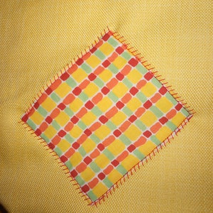 May include: A yellow fabric with a square patch of red, orange, and green checkered fabric sewn on top. The checkered fabric has a red zig-zag stitch around the edges.