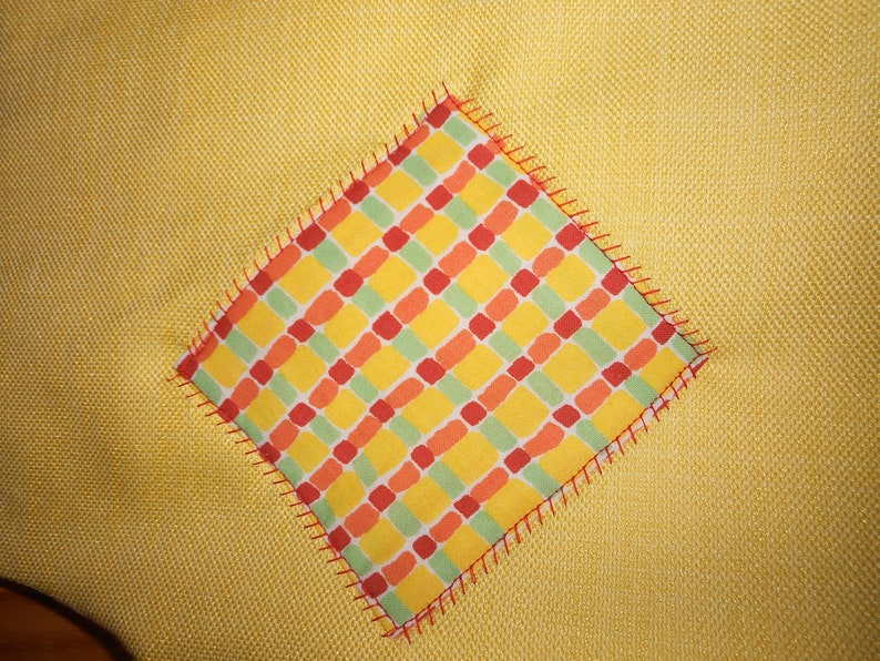 May include: A yellow fabric with a square patch of red, orange, and green checkered fabric sewn on top. The checkered fabric has a red zig-zag stitch around the edges.
