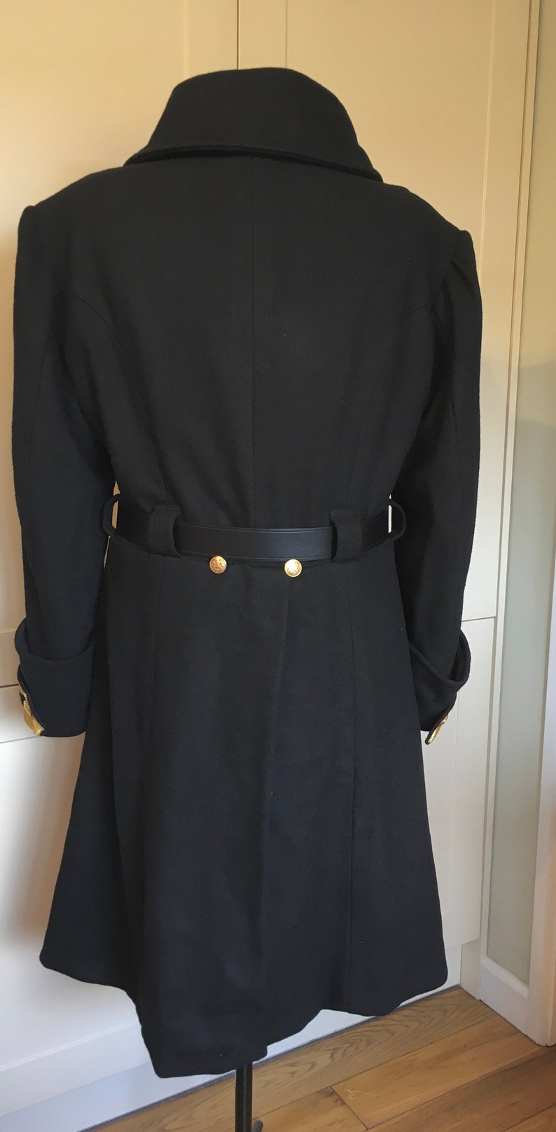 May include: A black wool coat with a black leather belt and gold buttons. The coat has a collar and long sleeves.