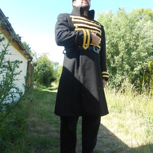 May include: A man wearing a black coat with gold trim and black pants. He is standing in a grassy area with trees in the background.