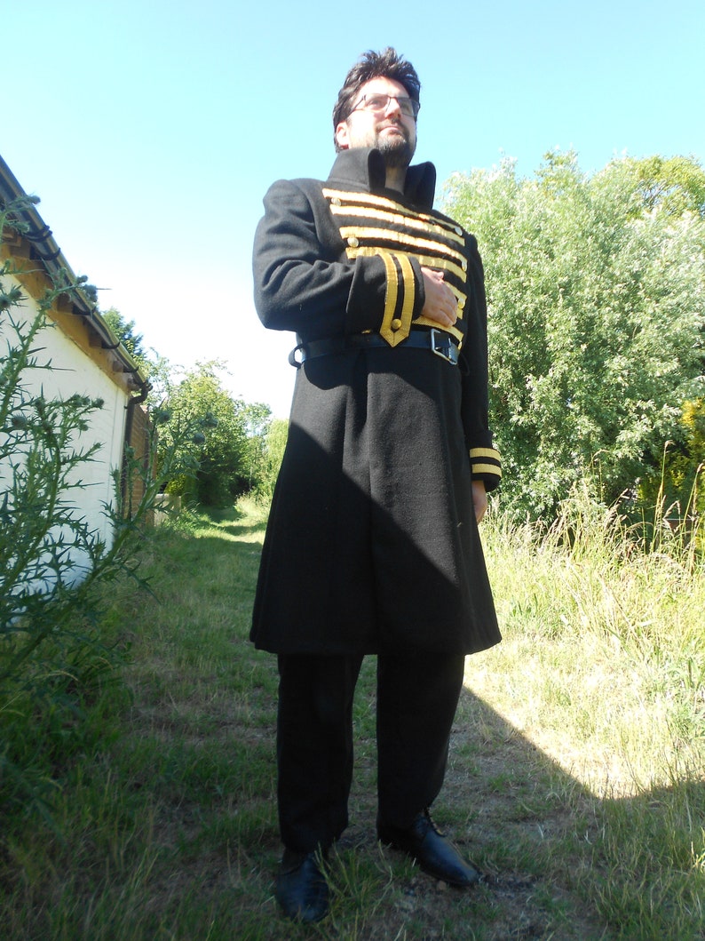 May include: A man wearing a black coat with gold trim and black pants. He is standing in a grassy area with trees in the background.