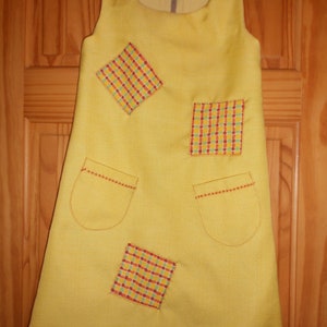 May include: A yellow sleeveless dress with four patchwork squares in red, yellow, and green checkered patterns. The dress has two pockets on the front.