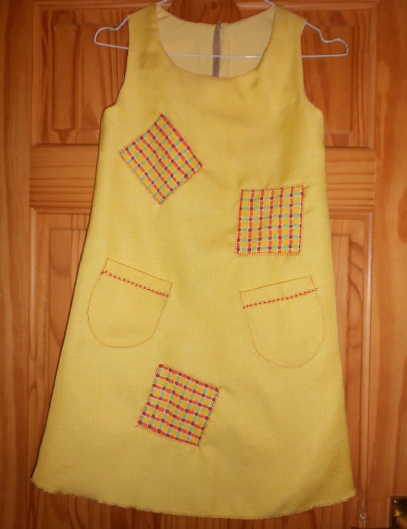May include: A yellow sleeveless dress with four patchwork squares in red, yellow, and green checkered patterns. The dress has two pockets on the front.