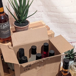 Puede incluir: Una caja de cart&oacute;n con una variedad de botellas de vidrio con etiquetas y tapas negras. Las botellas est&aacute;n llenas de un l&iacute;quido transparente. La caja est&aacute; sentada en una caja de madera. Hay una planta en una maceta de terracota a la izquierda de la caja.