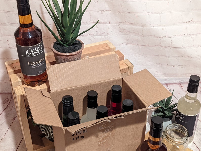 Puede incluir: Una caja de cart&oacute;n con una variedad de botellas de vidrio con etiquetas y tapas negras. Las botellas est&aacute;n llenas de un l&iacute;quido transparente. La caja est&aacute; sentada en una caja de madera. Hay una planta en una maceta de terracota a la izquierda de la caja.