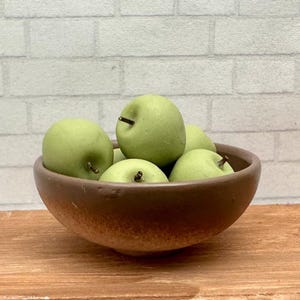 May include: A round, matte brown ceramic bowl filled with several smooth, light green artificial apples. Each apple features a small, dark brown stem protruding from the top. The bowl has a slightly textured, earthy finish with a darker base.