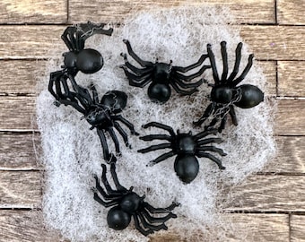 May include: Six black plastic spiders on a white spider web. The spiders have eight legs and a black body.