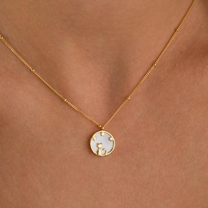 May include: A gold-colored necklace with a delicate chain and a round pendant. The pendant features a white background with a gold-colored design. The necklace is worn around the neck.