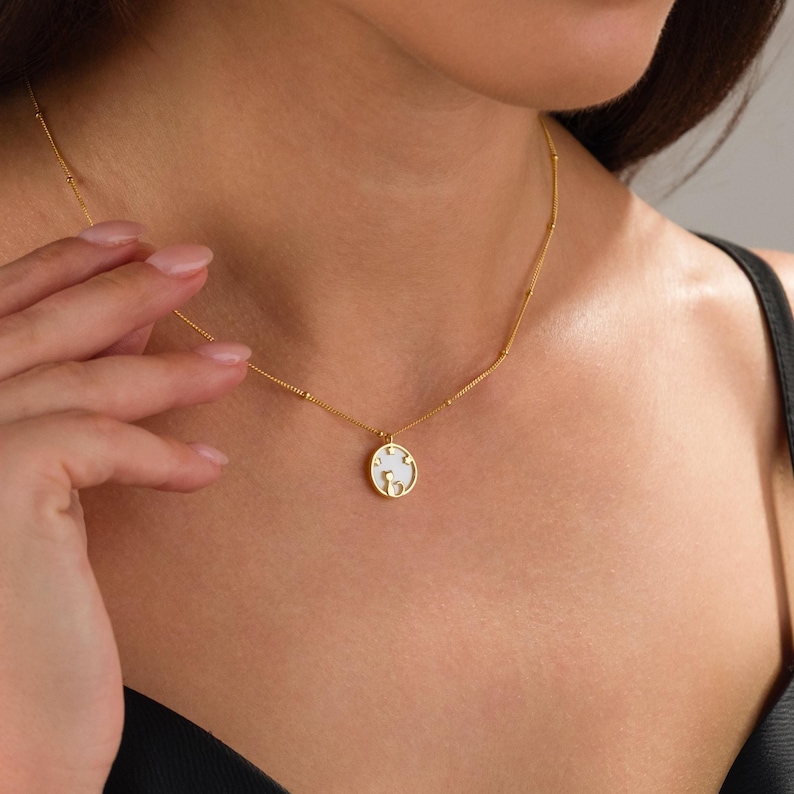 May include: A gold-colored necklace with a small, round pendant. The pendant features a white design and is suspended from a delicate chain. The necklace is worn around the neck.
