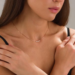 May include: A delicate rose gold necklace featuring a thin chain and a row of small, sparkling stones. The necklace is worn around the neck, showcasing its minimalist design and subtle elegance. The stones are set in a linear arrangement.