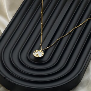 May include: A gold-colored necklace with a pendant featuring a cat silhouette and star accents. The oval pendant is white with a gold border. The necklace chain has small gold beads and a delicate design. The necklace is displayed on a black surface.