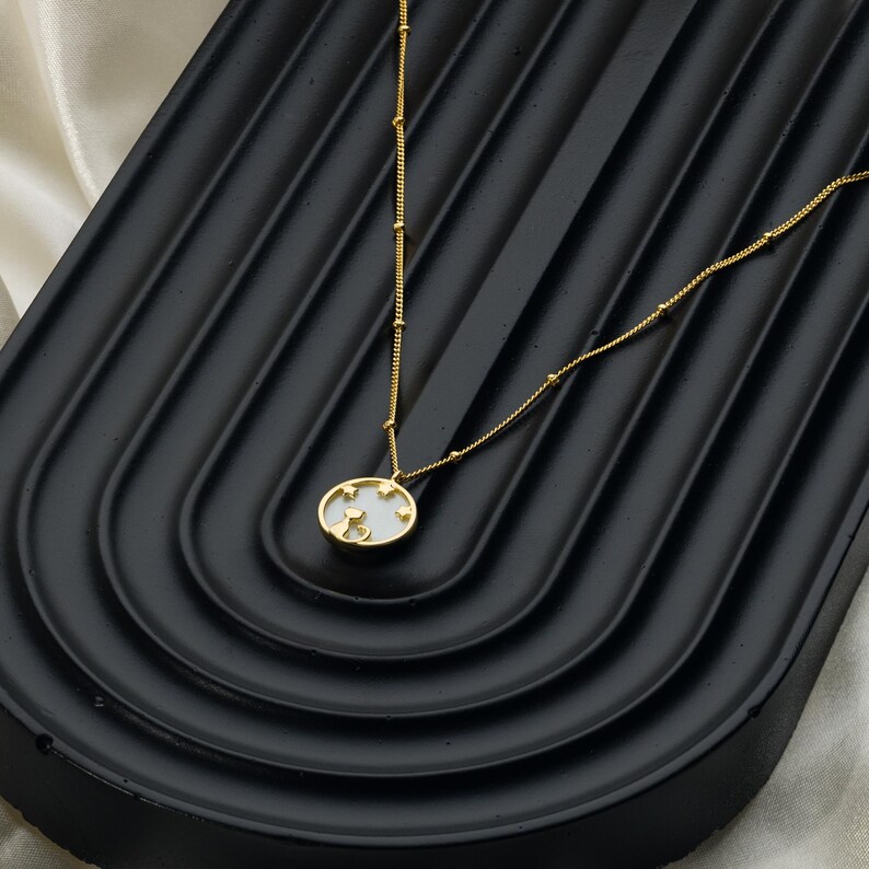 May include: A gold-colored necklace with a pendant featuring a cat silhouette and star accents. The oval pendant is white with a gold border. The necklace chain has small gold beads and a delicate design. The necklace is displayed on a black surface.
