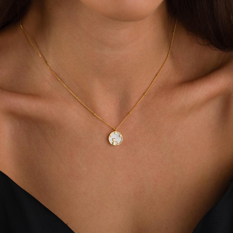 May include: A gold-colored necklace with a delicate chain and a round pendant. The pendant features a white background with a gold-colored design. The necklace is worn around the neck.