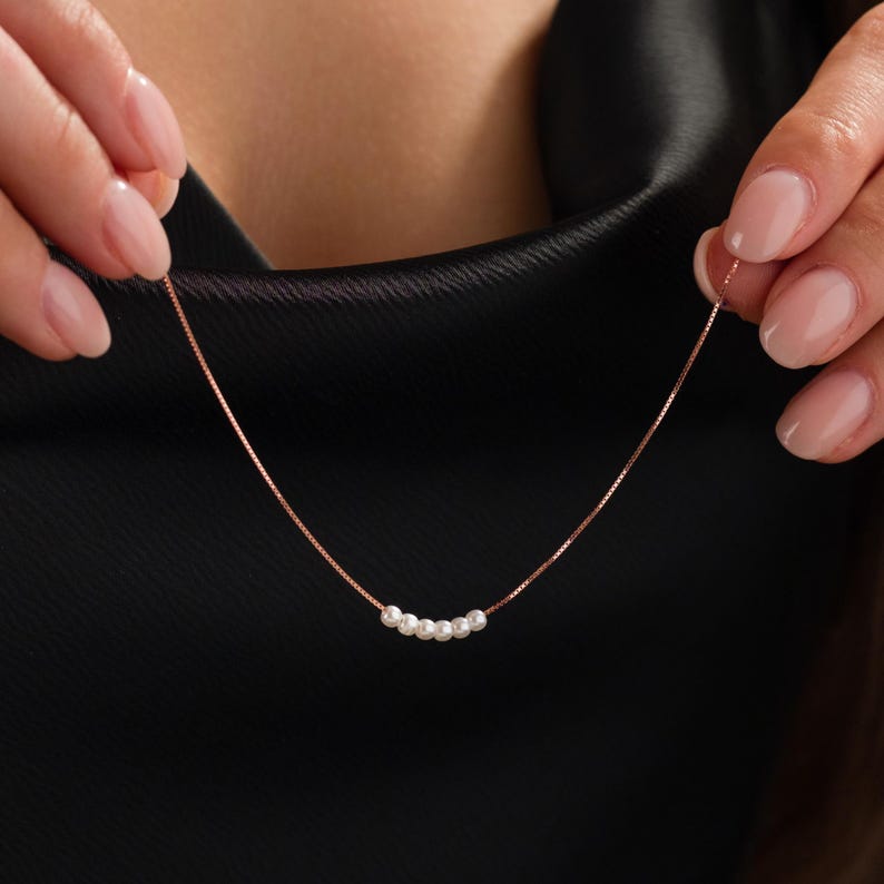 May include: A delicate rose gold necklace featuring a series of small, white pearls. The necklace has a fine chain and the pearls are evenly spaced. The background is a dark, solid color.