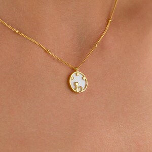 May include: Gold-colored necklace with a pendant featuring a cat silhouette and star accents. The pendant is round with a white background. The necklace is worn around the neck.