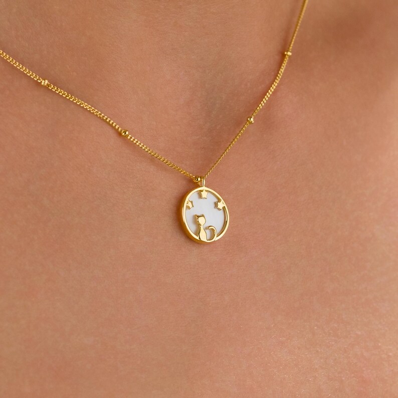 May include: Gold-colored necklace with a pendant featuring a cat silhouette and star accents. The pendant is round with a white background. The necklace is worn around the neck.