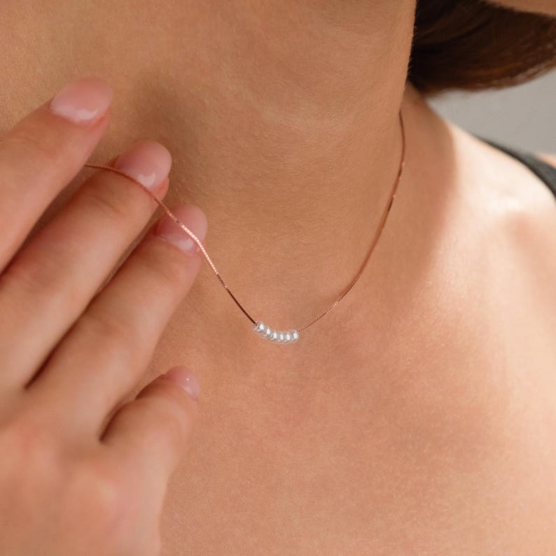 May include: A delicate necklace featuring a thin, rose gold chain. The necklace is adorned with a series of small, white pearls. The jewelry is displayed on a person's neck, with a hand gently touching the chain.