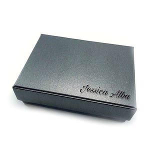 K&ouml;nnte beinhalten: Eine graue Geschenkbox mit dem Namen "Jessica Alba" auf der Oberseite.
