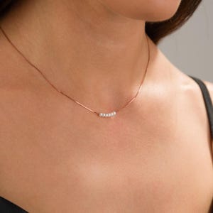 May include: A delicate necklace featuring a thin, rose gold chain. The necklace has a series of small, white pearls clustered together in the center. The jewelry is displayed against a neutral background.