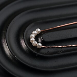 May include: A delicate necklace featuring a series of small, white pearls. The pearls are strung together in a curved arrangement, connected to a fine, rose-gold colored chain. The necklace rests on a black, textured surface.
