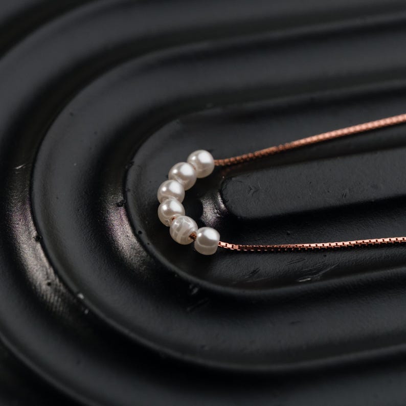 May include: A delicate necklace featuring a series of small, white pearls. The pearls are strung together in a curved arrangement, connected to a fine, rose-gold colored chain. The necklace rests on a black, textured surface.