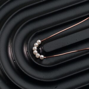 May include: A delicate necklace featuring a series of small, round, white pearls. The pearls are strung together in a curved line, connected by a thin, rose-gold colored chain. The necklace is displayed on a black, textured surface.