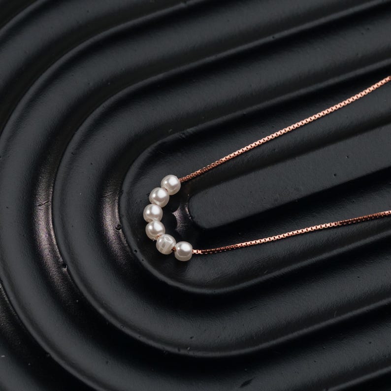 May include: A delicate necklace featuring a series of small, round, white pearls. The pearls are strung together in a curved line, connected by a thin, rose-gold colored chain. The necklace is displayed on a black, textured surface.
