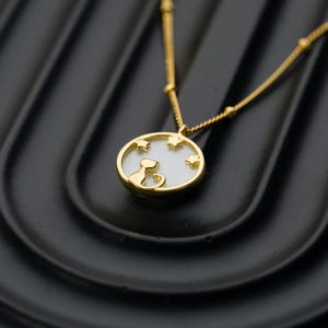 May include: Gold-toned necklace featuring a pendant with a cat silhouette and star cutouts. The pendant has a white background and a gold frame. The chain is delicate with small gold beads. The necklace is displayed on a black surface.
