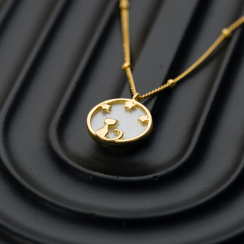 May include: Gold-toned necklace featuring a pendant with a cat silhouette and star cutouts. The pendant has a white background and a gold frame. The chain is delicate with small gold beads. The necklace is displayed on a black surface.